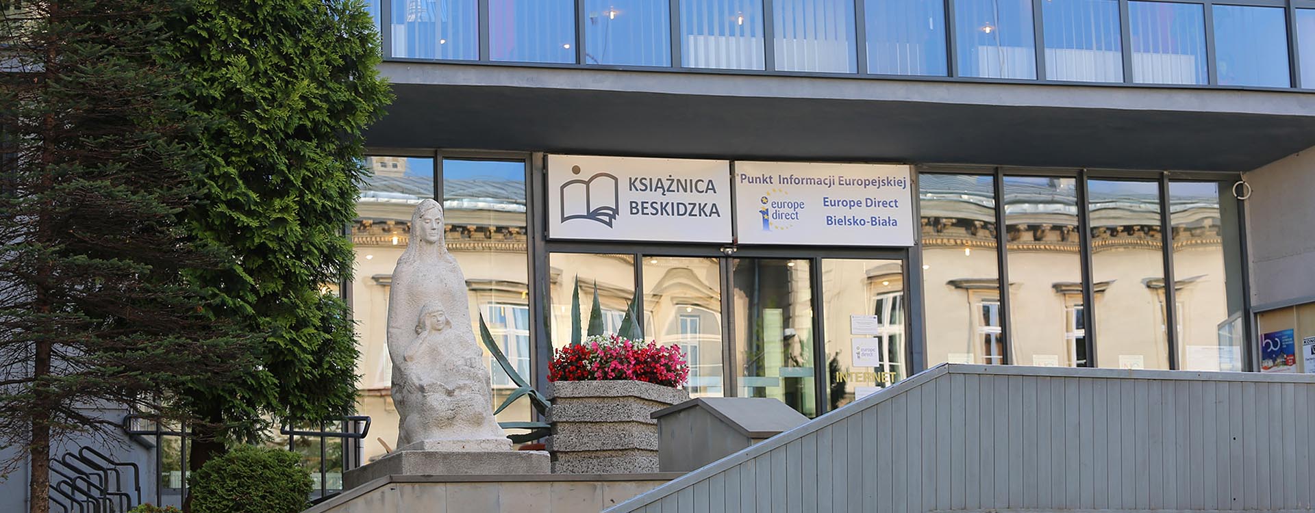 Widoczne jest oszkolone wejście do Książnicy od strony ulicy Słowackiego. Na górze, przy wejściu, po lewej stronie widać posąg oraz donicę z czerwonymi kwiatami. 